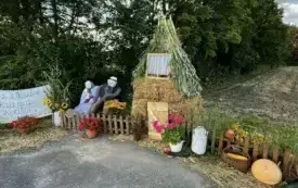 Sołectwo Watorowo. Witacz przedstawiajcy chatkę z płotem ozdobioną ważywami i wkiatami, przy kt&oacute;rej siedzi para