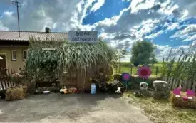 Sołectwo Bągart. Witacz przedstawiający chatkę przed kt&oacute;rą znajdują sie ważywa, kwiaty, biała kana, pies siedzący w budzie oraz dwie owce